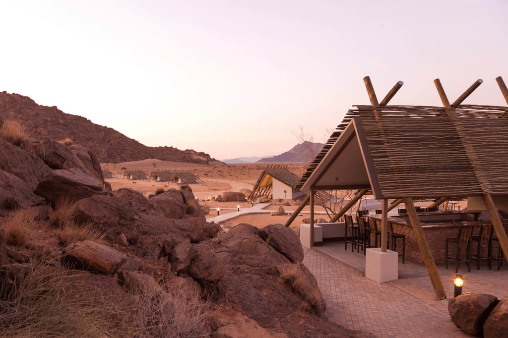 Wüstenlodge mit überdachtem Barbereich vor Felsen und roséfarbenem Abendhimmel
