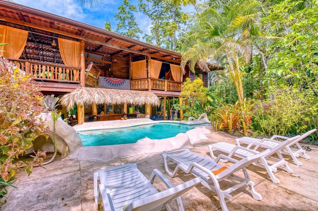 Zweistöckiges Holzhaus mit Veranda, Pool und Liegestühlen in dichtem tropischen Garten bei Tageslicht.