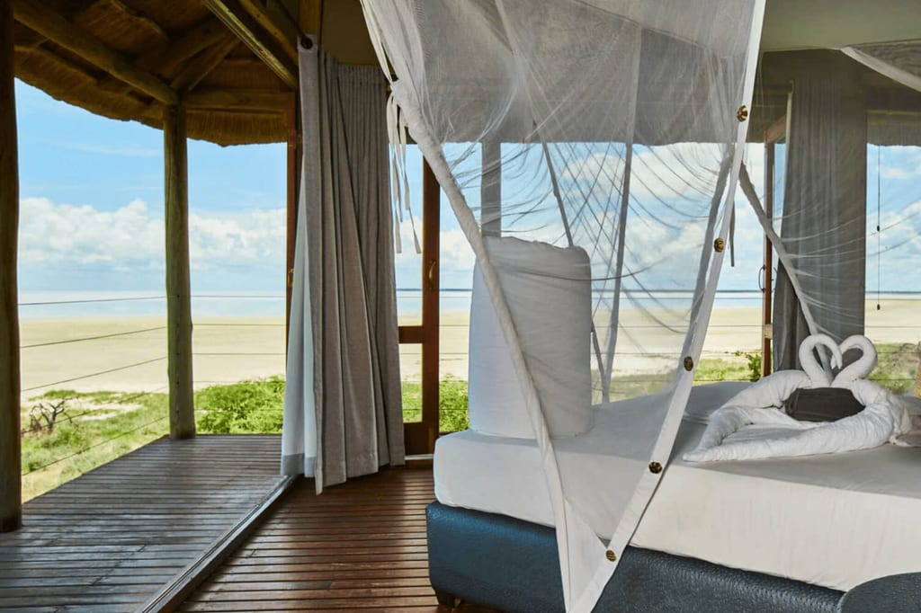 Offenes Strandbungalow-Zimmer mit Moskitonetz und Blick auf Sandstrand und Veranda bei Tageslicht.