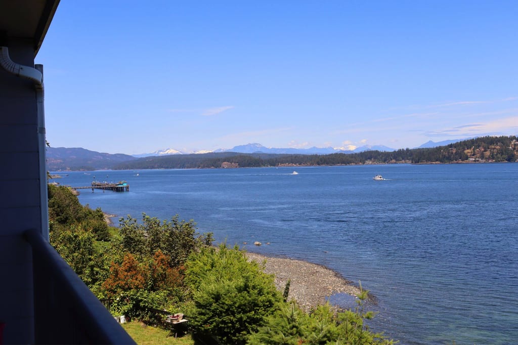 Blick von einem Balkon über eine ruhige Bucht mit Pier, Kiesstrand, Vegetation und schneebedeckten Bergen im Hintergrund.