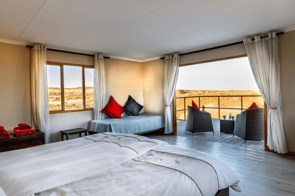 Helles Lodge-Schlafzimmer mit Doppelbett und offener Terrasse mit Sesseln und weitem, trockenem Ausblick.