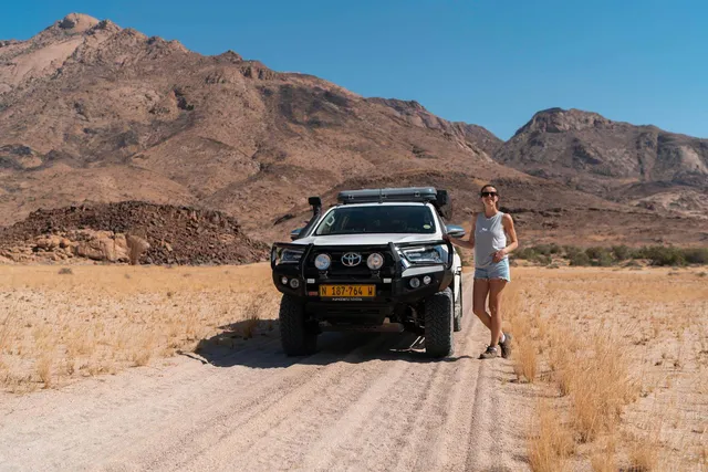 Frau neben Geländewagen auf Piste vor dem Brandberg in Namibia, sonnige trockene Landschaft