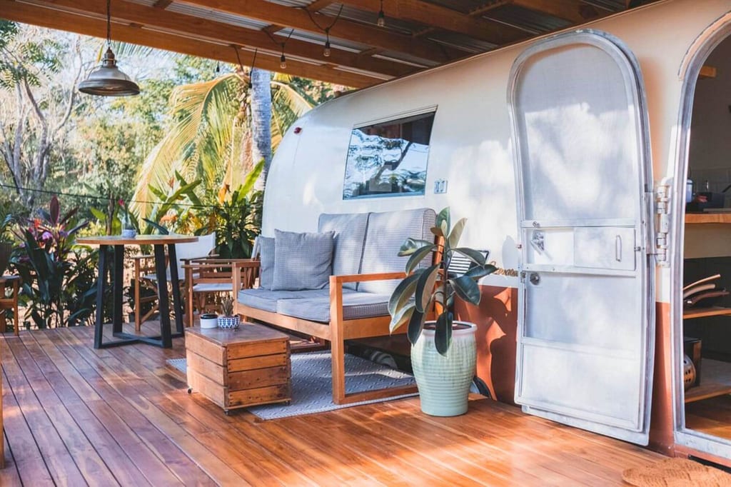 Wohnwagen auf Holzveranda mit Sofa, Holztisch und Topfpflanzen bei Sonnenschein