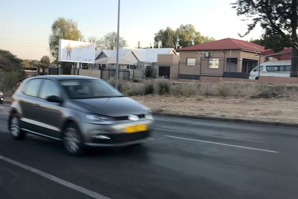 Fahrendes Auto auf Straße vor Wohnhäusern und Werbetafel in Rundu, Namibia