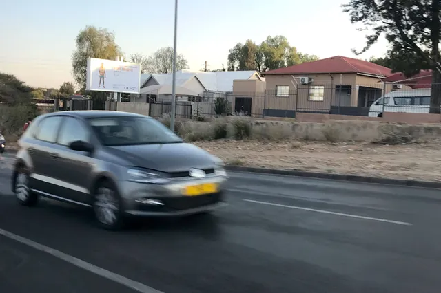 Fahrendes Auto auf Straße vor Wohnhäusern und Werbetafel in Rundu, Namibia