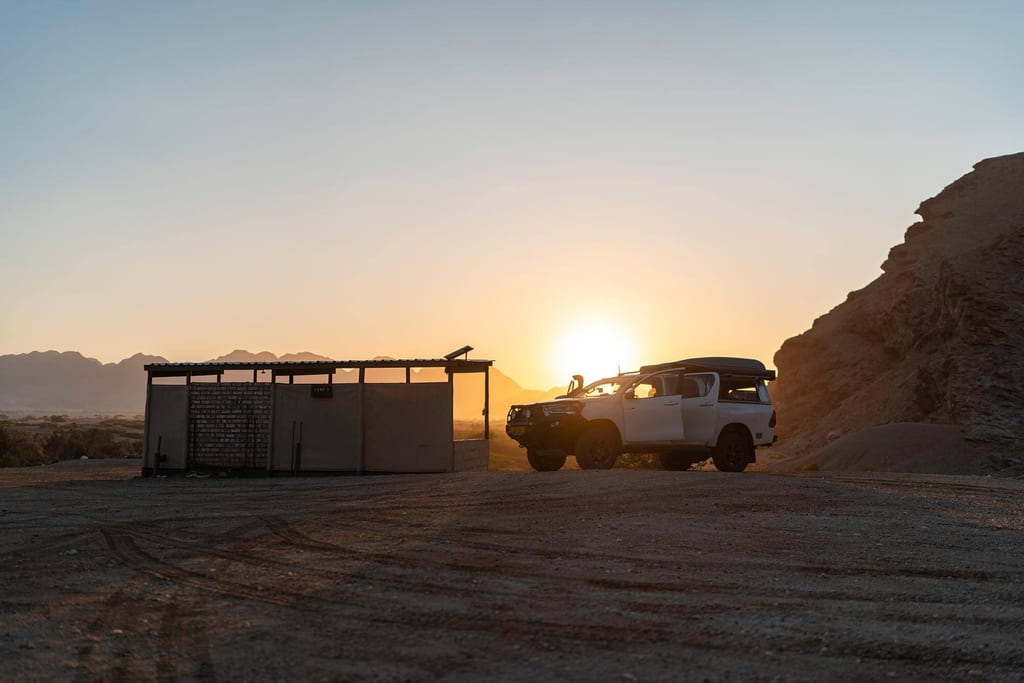 Weißer Geländewagen neben Unterstand in karger Wüstenlandschaft bei tief stehender Sonne.