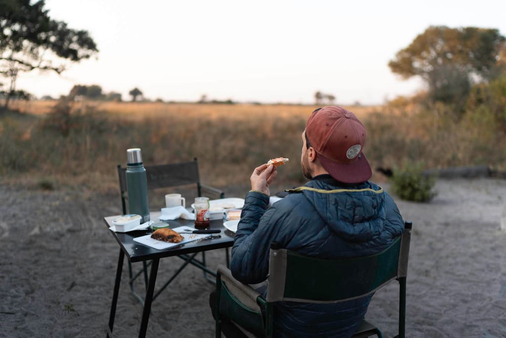 Person sitzt am Campingtisch und isst Brot, morgendliche Graslandschaft im Hintergrund.