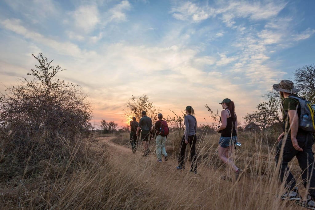 Sechs Personen wandern bei Sonnenuntergang auf einem Pfad durch trockenes Grasland mit Dornbüschen.