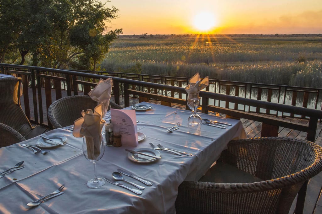 Gedeckter Tisch auf Holzterrasse mit Blick auf Schilf und Sonnenuntergang am Gewässer.