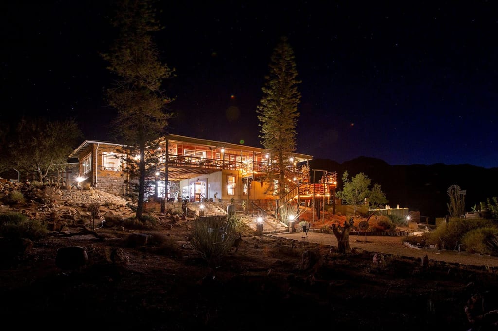 Beleuchtete Lodge bei Nacht vor klarem Sternenhimmel auf trockenem, felsigem Gelände.