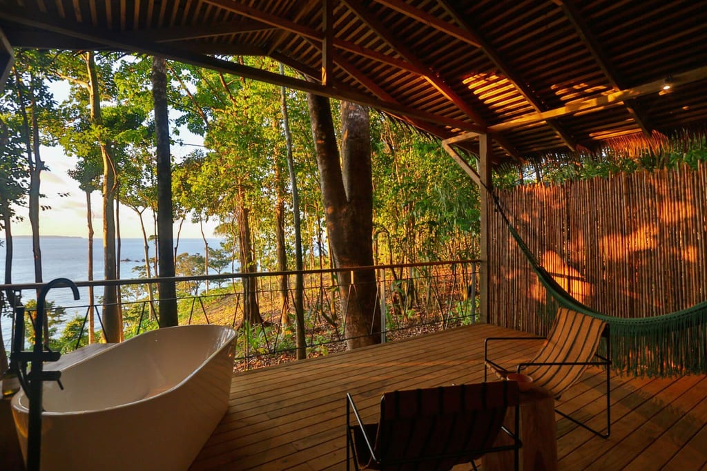 Überdachte Holzveranda mit Badewanne und Hängematte, Blick durch Bäume aufs Meer im warmen Abendlicht.