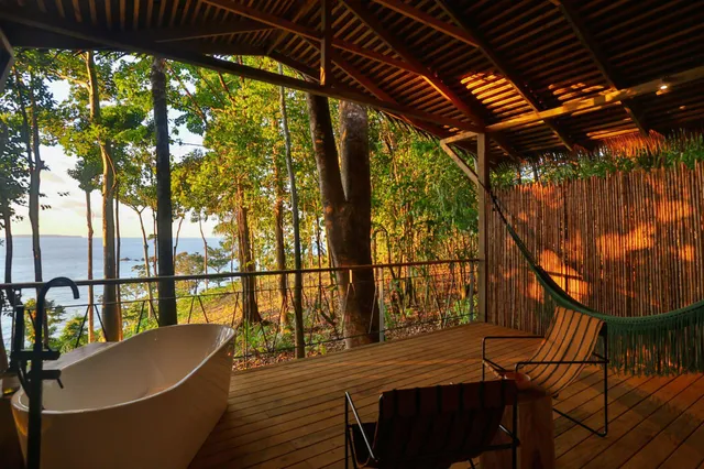 Überdachte Holzveranda mit Badewanne und Hängematte, Blick durch Bäume aufs Meer im warmen Abendlicht.