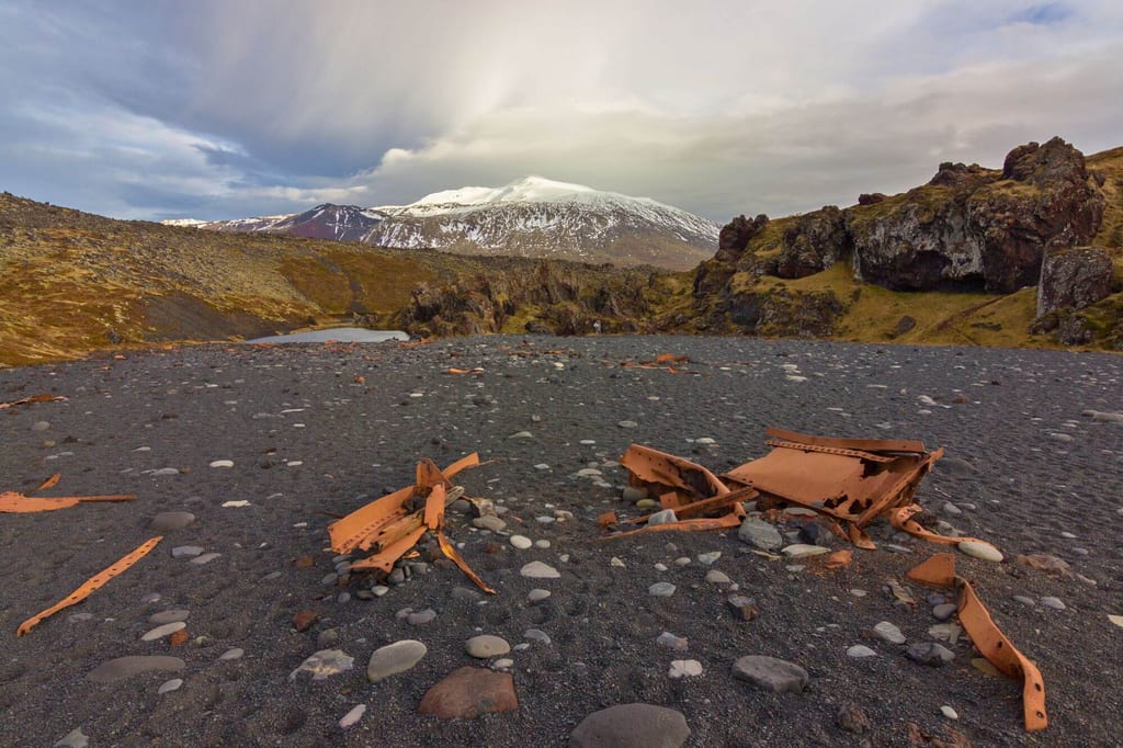 Schwarzer Kiesstrand mit verrosteten Metalltrümmern, moosige Felsen und schneebedeckter Berg unter Wolken.