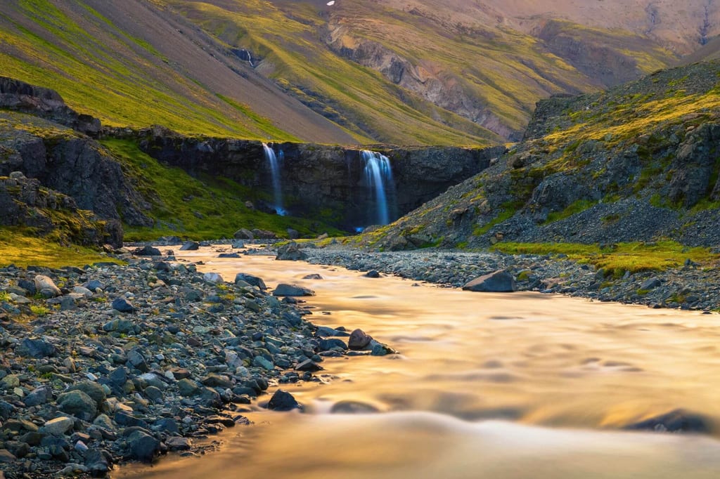 Fluss mit glattem Wasser läuft auf einen zweigeteilten Wasserfall in einem grünen Gebirgstal im warmen Abendlicht.