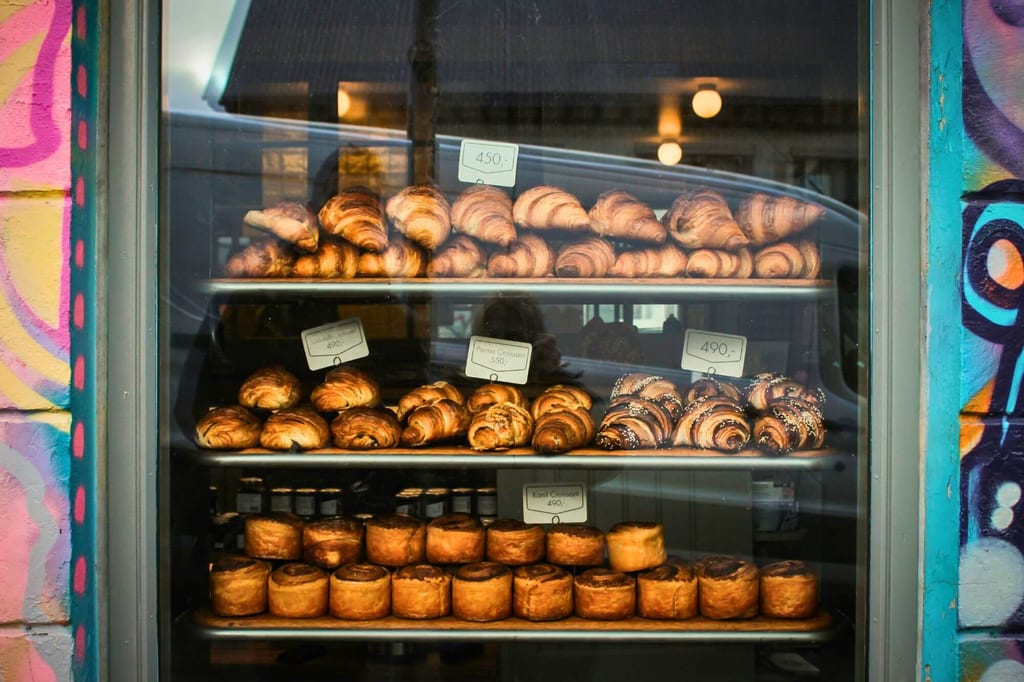 Schaufenster einer Bäckerei mit Croissants und Gebäck auf drei Regalböden, Straßenreflexionen im Glas.