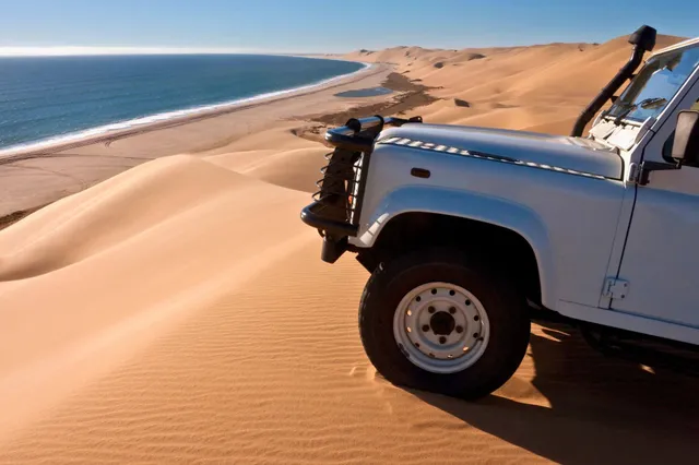 Geländewagen auf Sanddüne mit Blick auf Küste und ruhiges Meer im hellen Tageslicht.