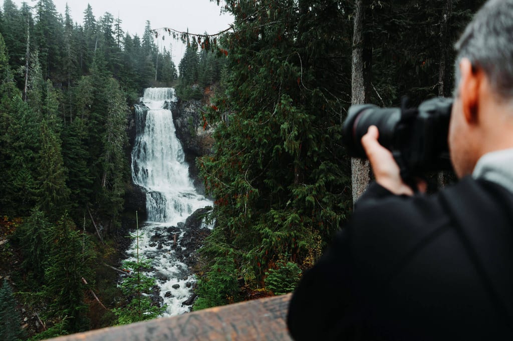 Fotograf auf Holzgeländer fotografiert mehrstufigen Wasserfall im dichten Nadelwald bei bewölktem Himmel.