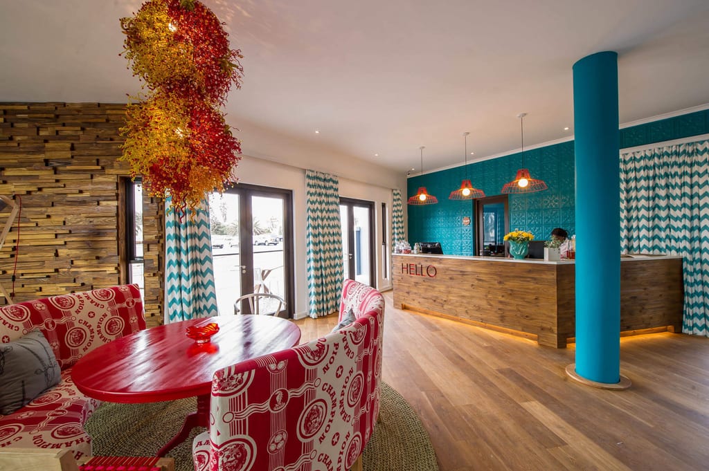 Bunte Hotel-Lobby mit rotem Tisch, gemusterter Sitzbank und Holz-Reseption bei Tageslicht.