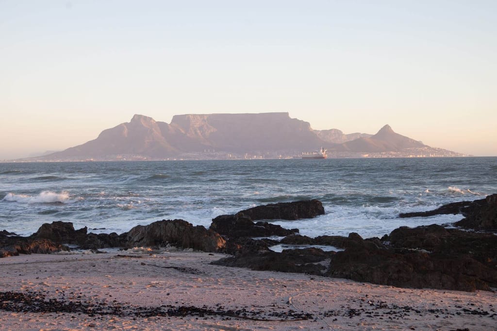 Blick vom felsigen Strand über das Meer zu einem flachen Bergmassiv im warmen Dämmerlicht