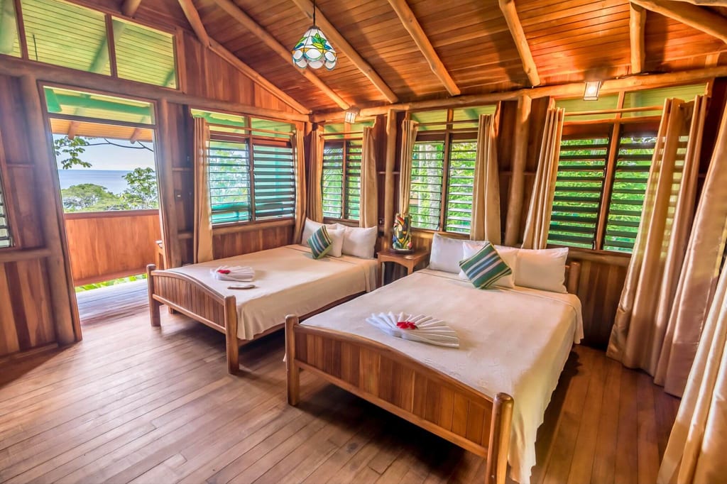 Helles Holzschlafzimmer in einem Bungalow mit zwei Betten, geöffneten Fenstern und Meerblick.