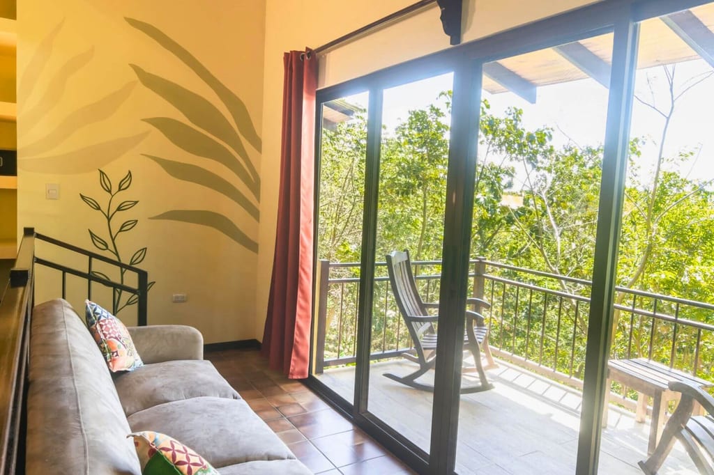 Helles Wohnzimmer mit Sofa und Balkon mit Schaukelstuhl und Blick auf grünes Baumgrün.