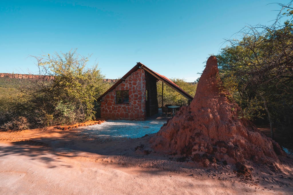 Kleines Steinhaus mit großem Termitenhügel vor trockenem Dornbusch bei klarem Himmel