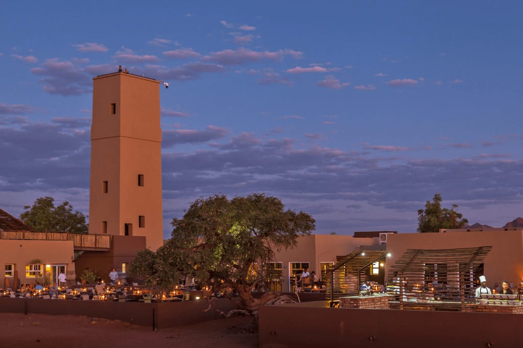 Abendliche Szene: Turm und beleuchteter Innenhof mit Gästen und Kerzen unter violettem Himmel
