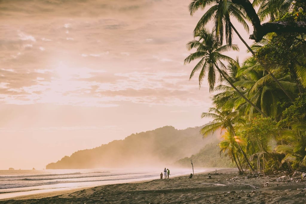 Tropischer Strand im Corcovado Nationalpark mit Palmen, Nebel und Menschen als Silhouetten bei goldener Dämmerung.