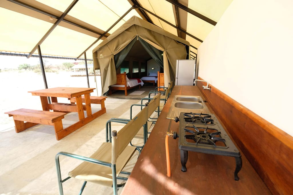 Offene Safari-Zeltunterkunft mit Außenküche, Holztisch und sichtbaren Betten in hellem Tageslicht.