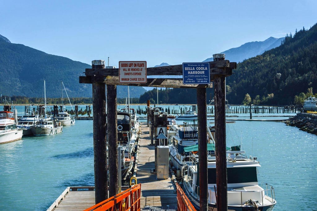 Blick auf Holzsteg und festgemachte Boote im Bella Coola Harbour, umgeben von Bergen und klarem Himmel.