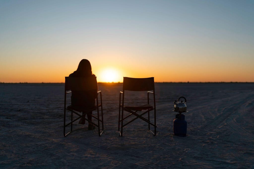 Person sitzt in Campingsessel als Silhouette, daneben leerer Stuhl und Teekessel, Sonnenuntergang über weiter Fläche