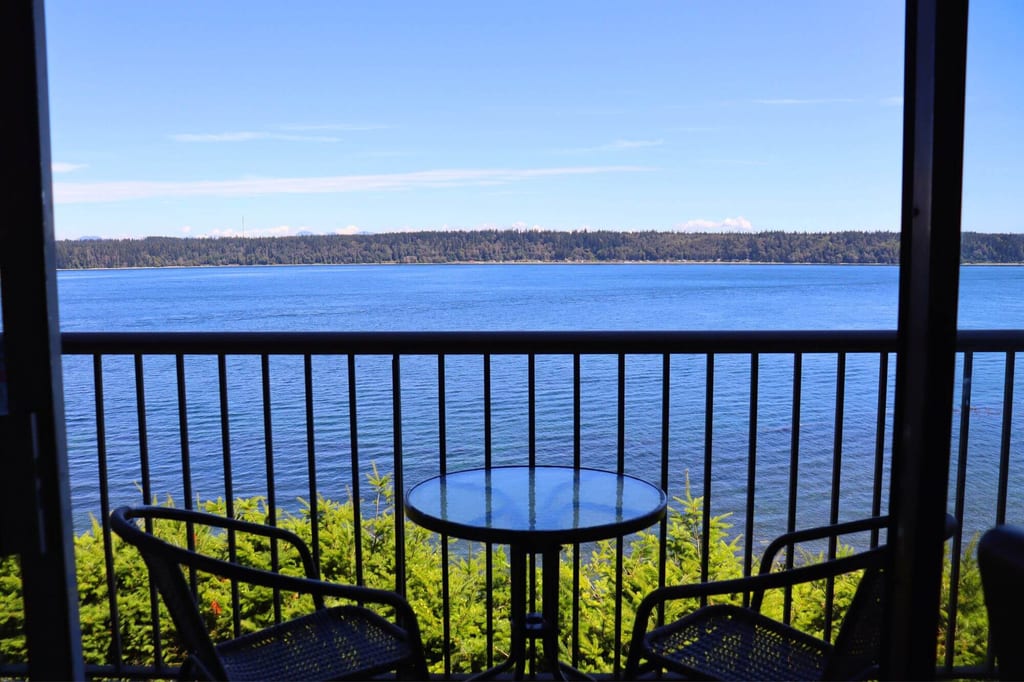 Balkon mit Tisch und zwei Stühlen blickt auf ruhiges blaues Wasser und bewaldetes Ufer bei klarem Himmel.