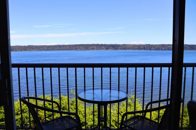 Balkon mit Tisch und zwei Stühlen blickt auf ruhiges blaues Wasser und bewaldetes Ufer bei klarem Himmel.