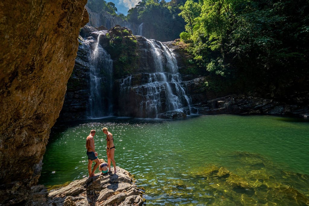 Wasserfall mit grünem Pool, drei Personen in Badebekleidung auf einem Felsen am Ufer.