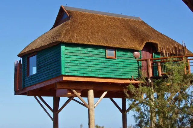 Erhöhte grüne Holzhütte mit Strohdach auf Pfosten vor blauem Himmel