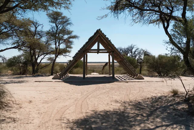 Dreieckiges Holzshelter auf sandigem Boden in trockener Savannenlandschaft, klarer Himmel