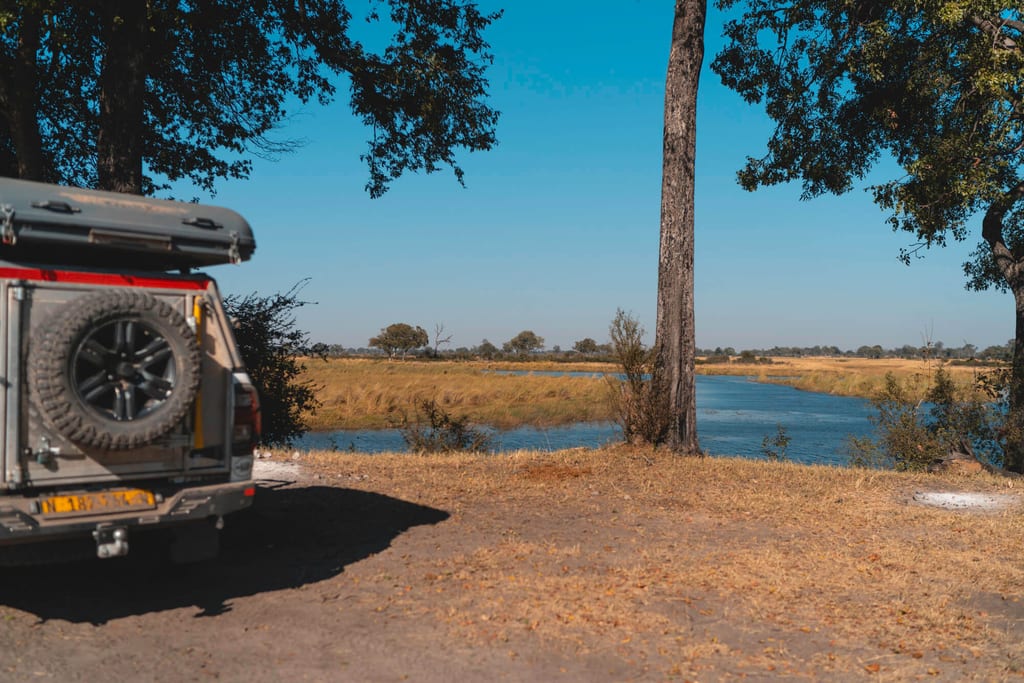 Geländewagen am Ufer eines ruhigen Flussarms, weite Aue und klarer blauer Himmel