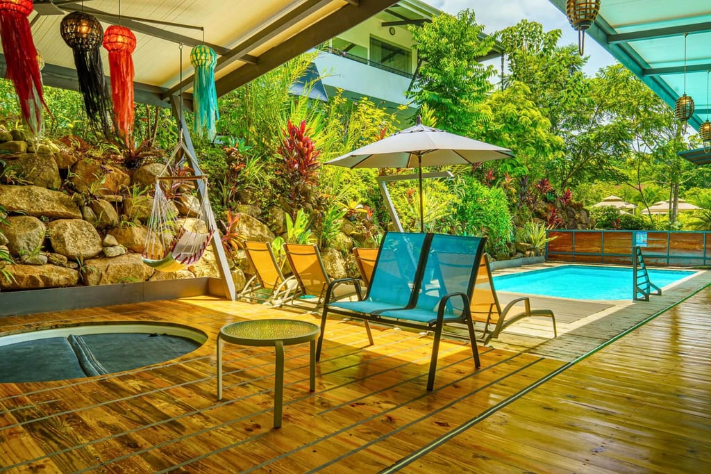 Überdachte Poolterrasse mit Holzdeck, Liegestühlen, Hängesesseln und dichter tropischer Bepflanzung.