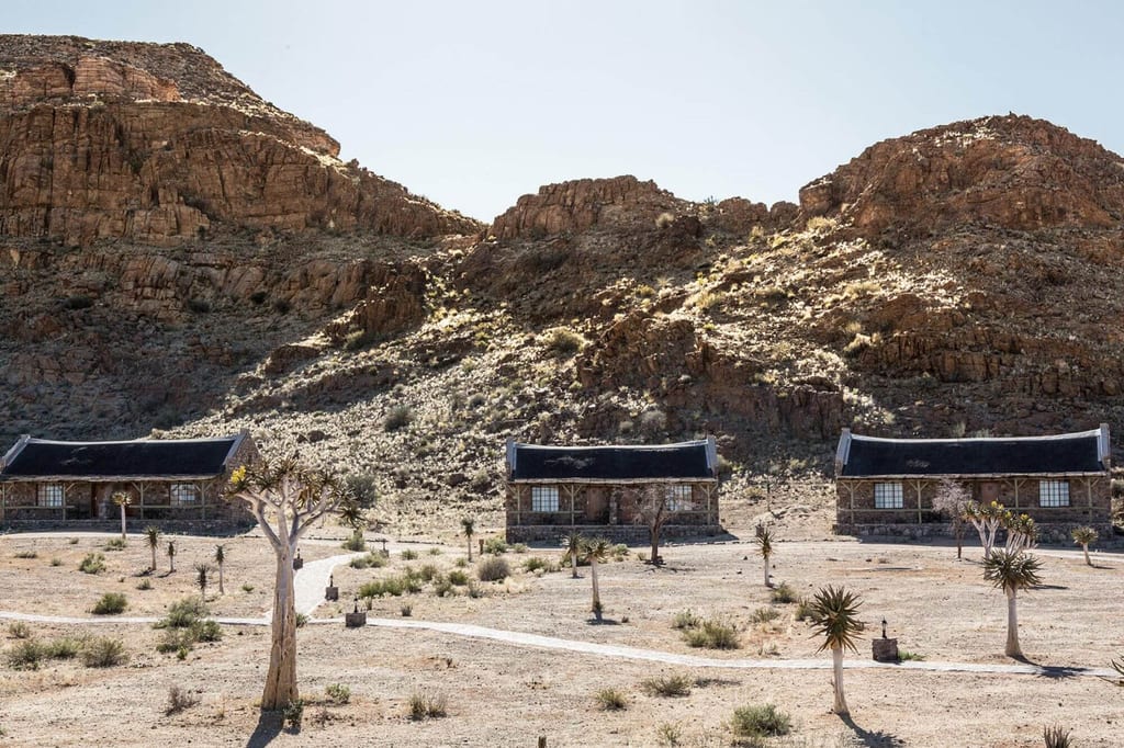 Drei Lodge-Häuser vor trockenen Felsrücken und vereinzelten Bäumen im harten Mittagslicht.