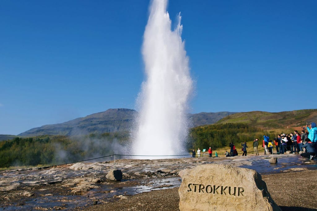 Hohe Geysir-Wasserfontäne vor blauem Himmel, Besucher beobachten, Findling mit Inschrift im Vordergrund.