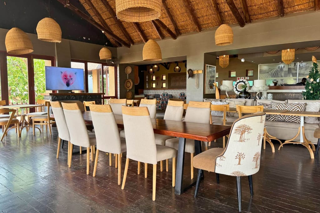 Offene Lodge-Lounge mit langem Holztisch, gepolsterten Stühlen und Strohdach bei Tageslicht.