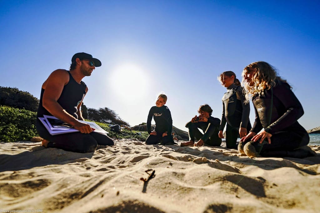 Fünf Personen in Neoprenanzügen sitzen im Gegenlicht auf einem Sandstrand, eine Person hält Papiere.