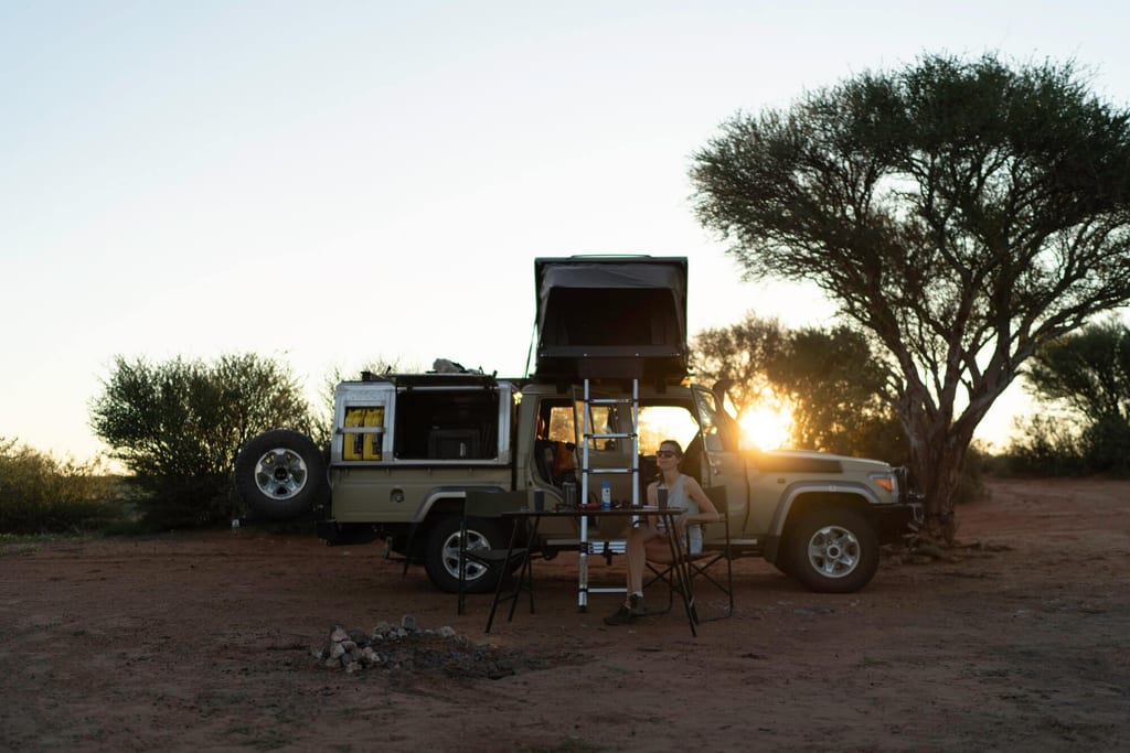 Geländewagen mit Dachzelt und Person am Campingtisch bei Sonnenuntergang in trockener Landschaft