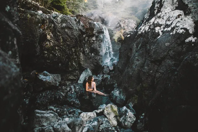 Person sitzt in einem Felsbecken vor einem schmalen Wasserfall in einer felsigen Schlucht.