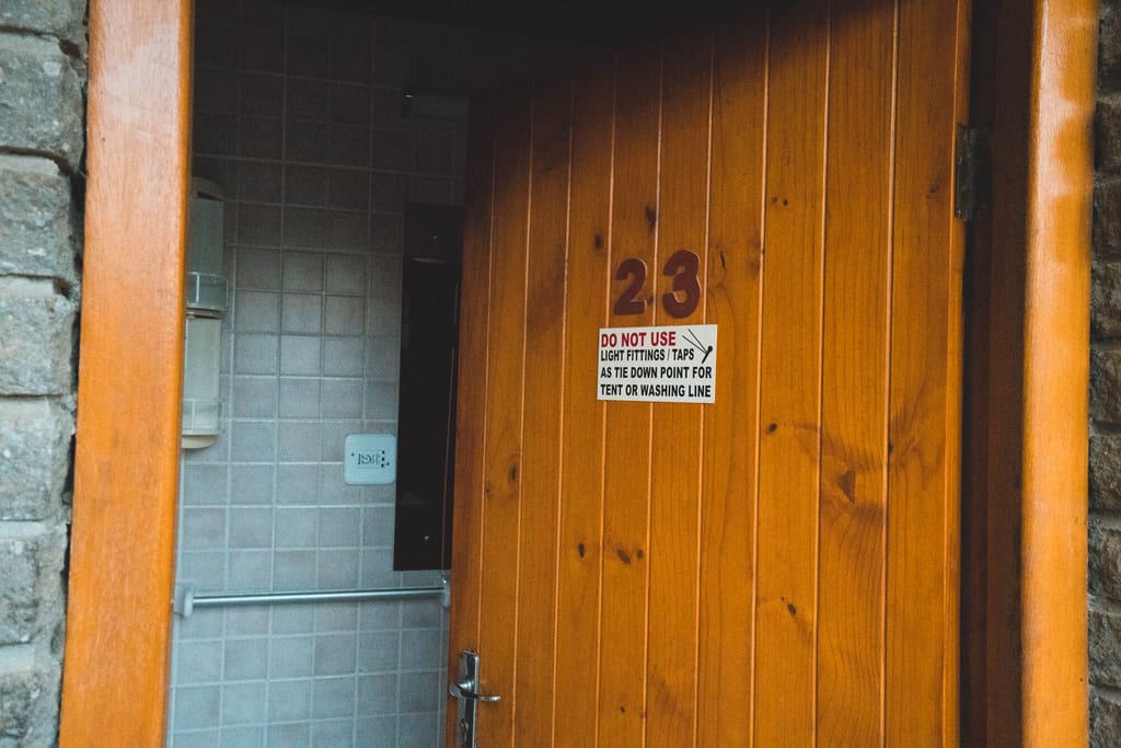 Offenstehende Holztür mit Nummer 23 und Warnaufkleber, Blick ins geflieste Innere.