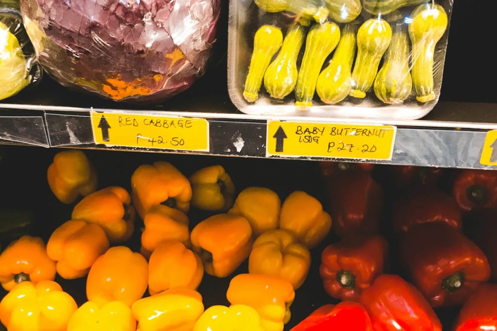 Nahaufnahme eines Supermarktregals mit gelben und roten Paprika, Rotkohl und verpackten kleinen Butternut-Kürbissen.