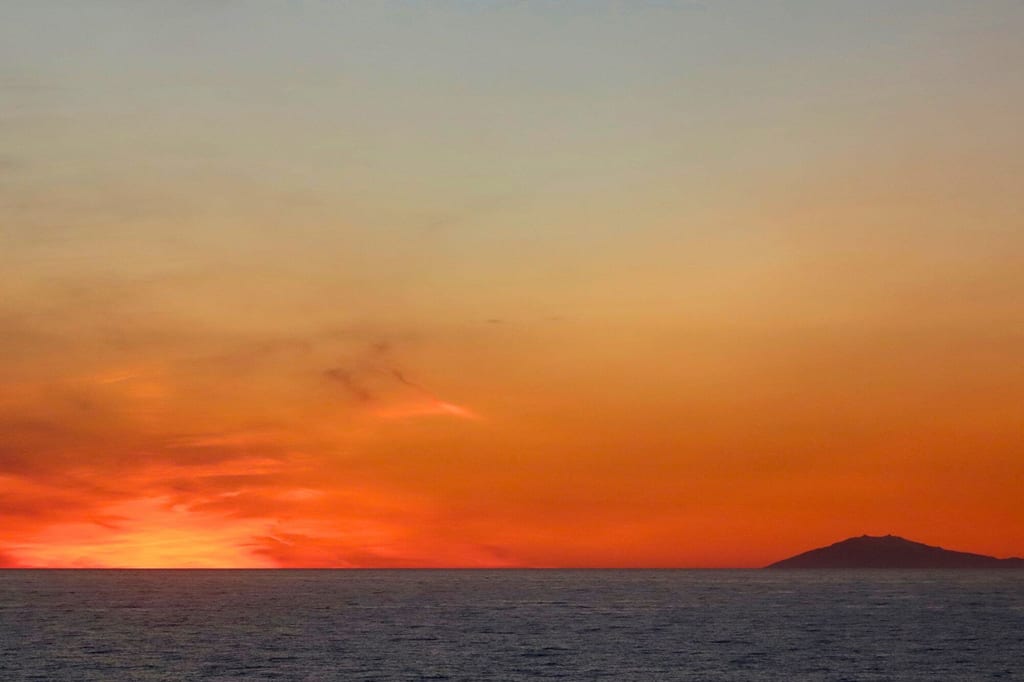Ruhevolles Meer bei intensiv orangefarbenem Sonnenuntergang mit Insel-Silhouette am Horizont.