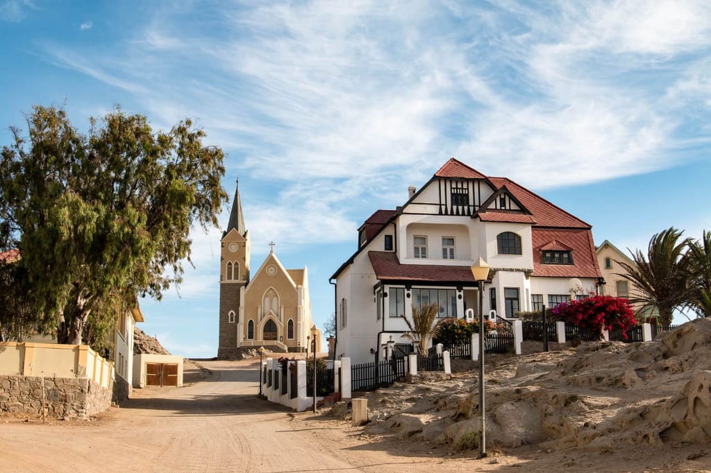 Straße in Lüderitz mit Kirche im Hintergrund und weißem Haus mit rotem Dach, sonniger Tag