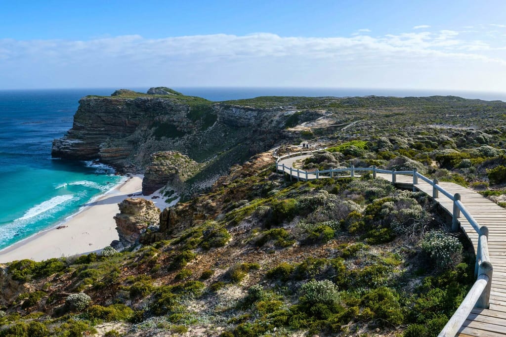 Hölzerner Bohlenweg über Küstenvegetation zu Klippen und sandigem Strand mit türkisfarbenem Meer