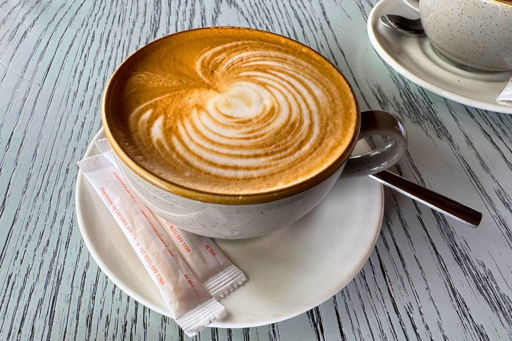 Nahaufnahme einer Tasse Cappuccino mit spiralförmigem Milchschaum auf Untertasse, Zuckersticks und Löffel auf Holztisch.